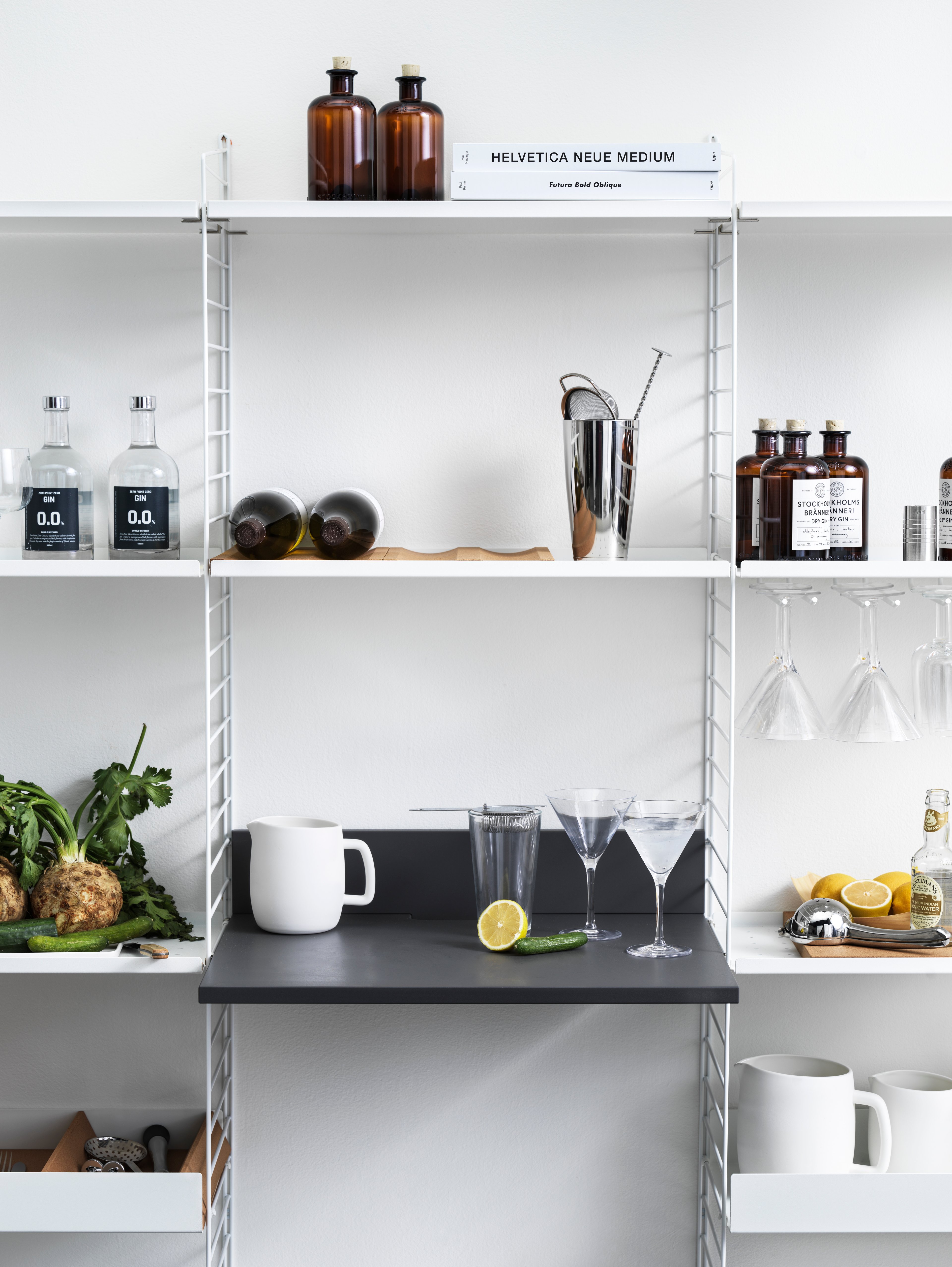 String shelf with floor panels from String System. Panels, metal shelves with low edge, metal shelves with high edge, and hanger racks white. Media shelf in anthracite grey, bottle tray in cork, divider in cork, and underlay in cork. This shelving combination is perfect in a kitchen or living room where the media shelf can be used as a drink table.