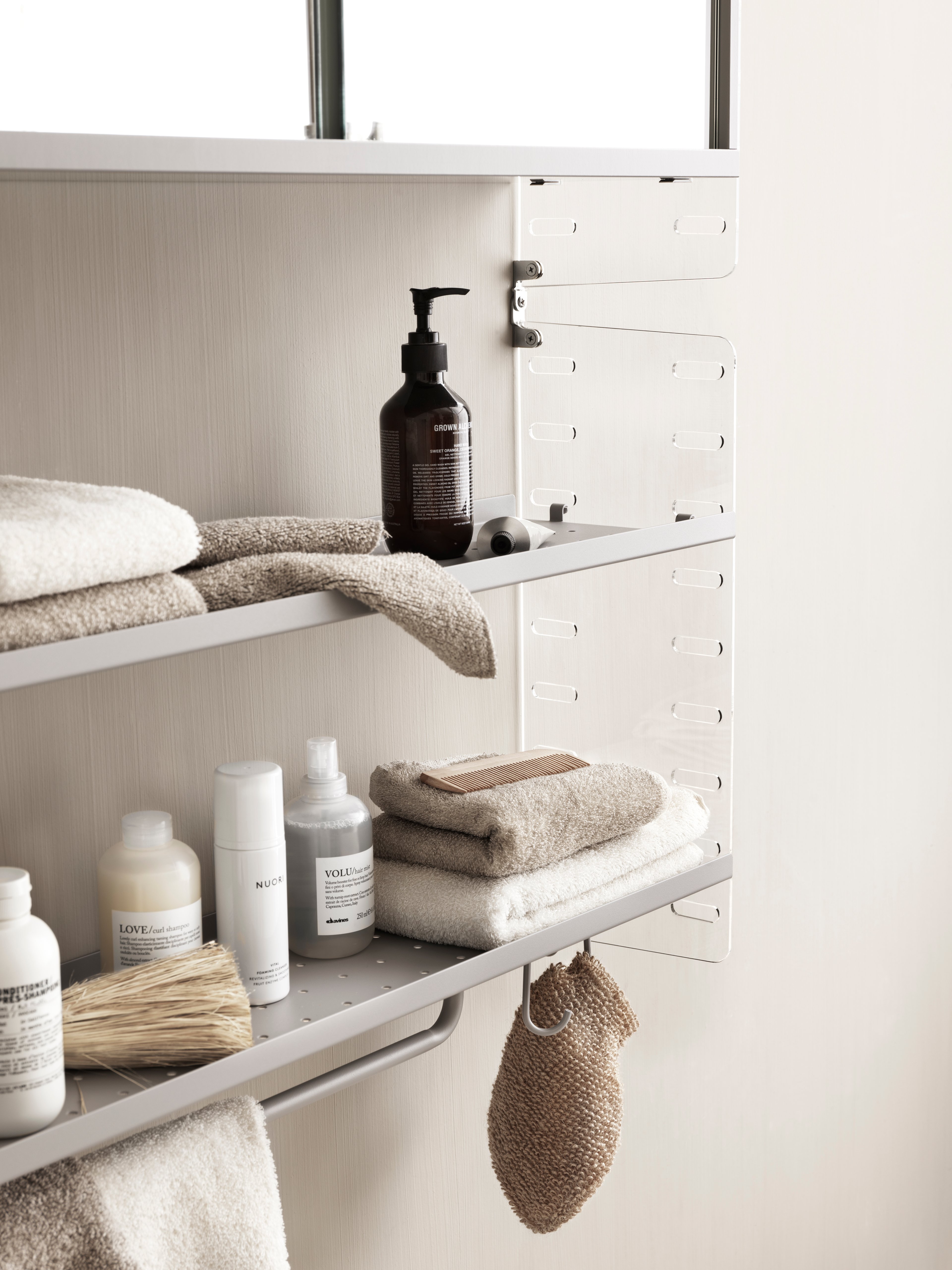 Solución de baño montada en pared de String. Paneles de pared en Perspex transparente, estantes metálicos de borde bajo y cabinet con puertas correderas, barra de colgar y ganchos en gris.