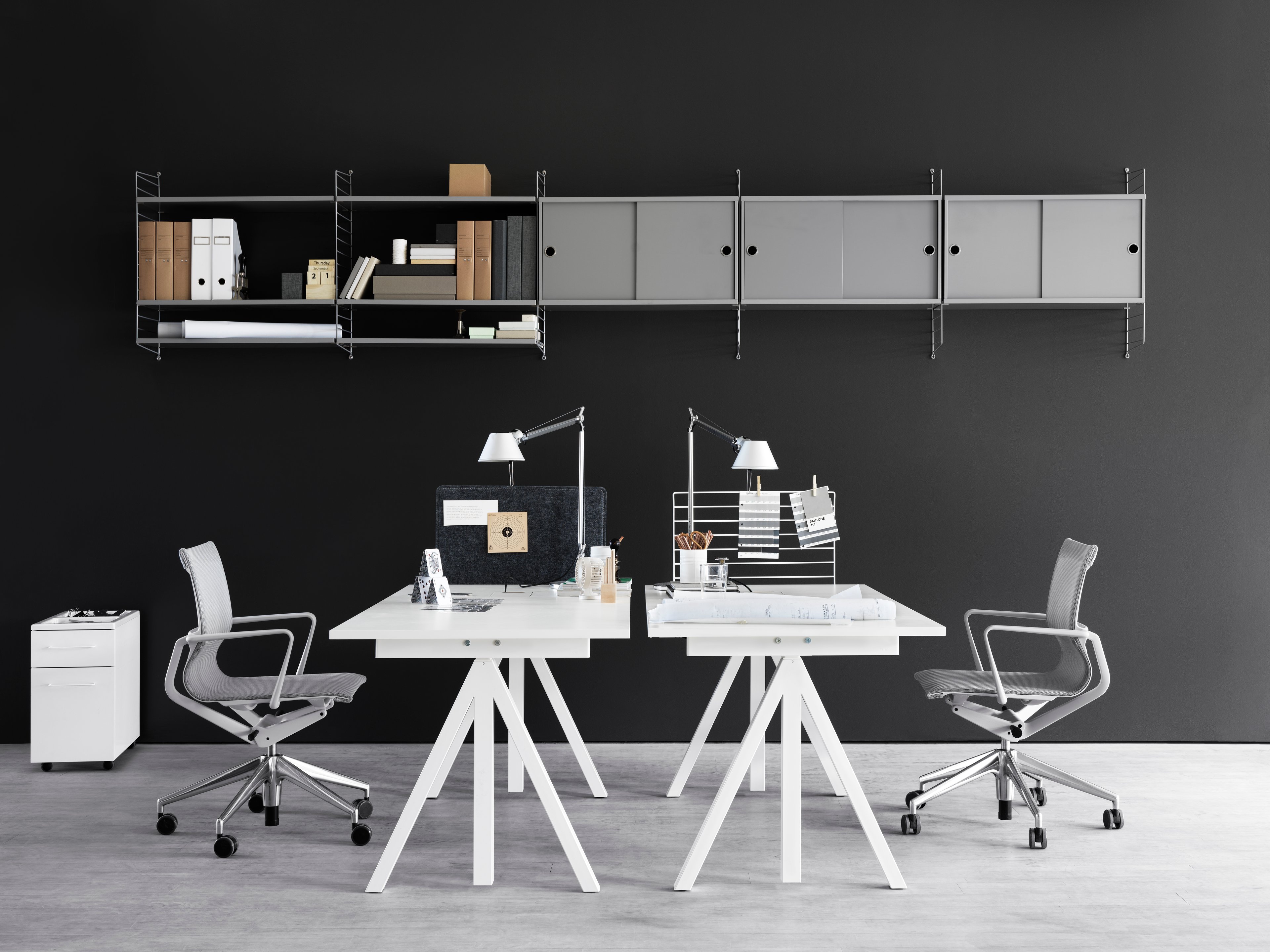 Wall mounted workspace solution from String. Height-adjustable work desk in white. Mobile storage in white. Wire screen in white. Fabric sleeve in grey felt. Freestanding shelf. Wall panels in grey. Shelves and cabinets with sliding doors in grey.